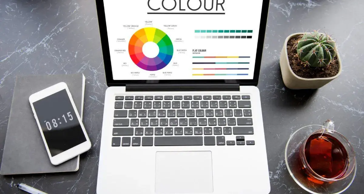 Na ekranie laptopa koło barw i palety kolorów, pomocne przy wyborze między rgb czy cmyk do druku. Obok telefon, notes i kaktus.