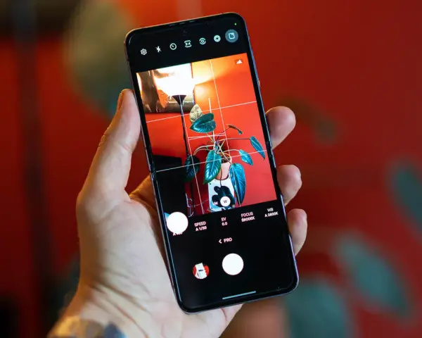 Dłoń trzyma smartfon z włączonym trybem PRO aparatu. Na ekranie widoczny jest kadr z rośliną w doniczce i lampą.