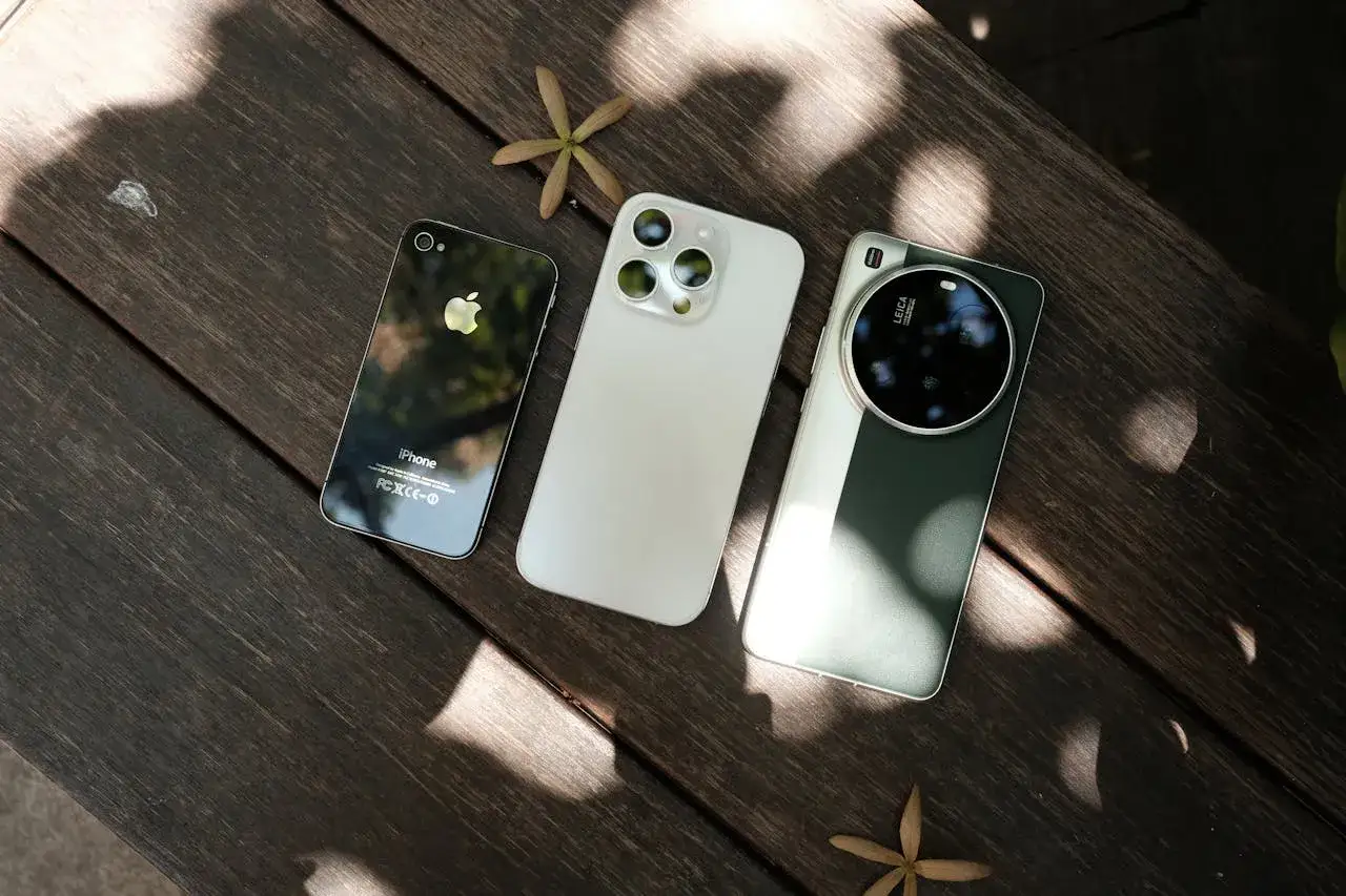 Trzy telefony na drewnianym stole: stary iPhone, biały smartfon i zielony telefon do zdjęć do 2000 z aparatem Leica.
