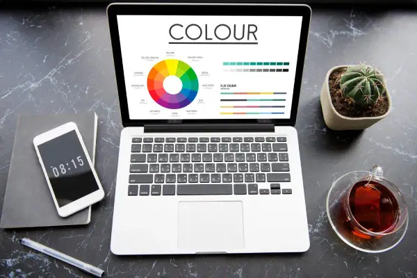 Na ekranie laptopa koło barw i palety kolorów, pomocne przy wyborze między rgb czy cmyk do druku. Obok telefon, notes i kaktus.